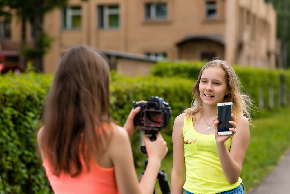 Two girls are bloggers. Young journalists. Describes advantages of phone. Make a broadcast on the Internet. Record vlog and blog subscribers. Record video lesson for Internet. Use camera with tripod.